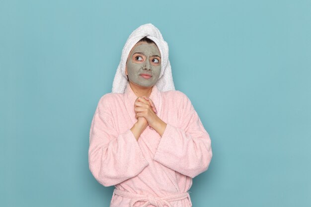 Вид спереди молодая женщина в розовом халате с маской на лице, мечтающая на синей стене, чистка душа, крем для ухода за собой