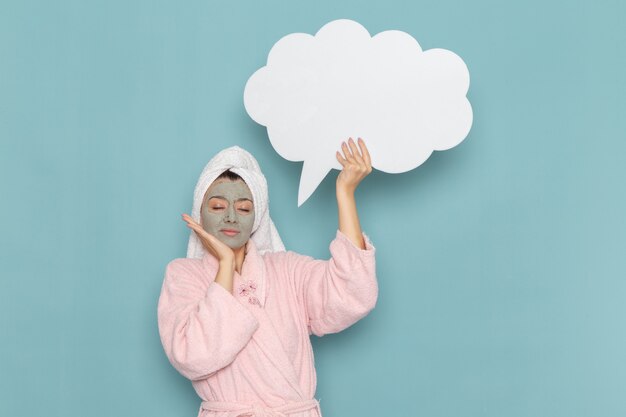 Front view young female in pink bathrobe with mask on her face holding big white sign on blue wall shower cleaning beauty selfcare cream