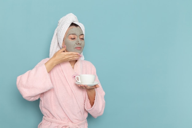 ピンクのバスローブを着た若い女性の正面図青い壁にコーヒーを保持している彼女の顔にマスクと美容セルフケアクリーム