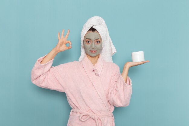 Front view young female in pink bathrobe with mask on her face holding cream on blue desk shower cleaning beauty self-care cream