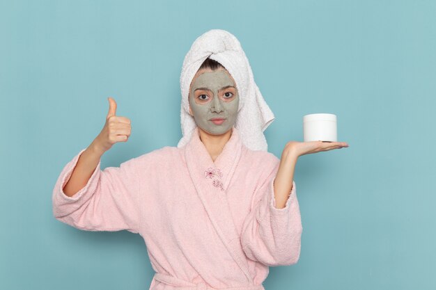 Front view young female in pink bathrobe with mask on her face holding cream on blue wall shower cleaning beauty selfcare cream