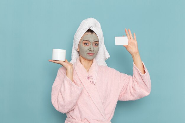 Front view young female in pink bathrobe with mask on her face holding cream on the light blue wall shower cleaning beauty self-care cream