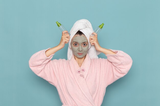 青い壁のシャワーのクリーニング美容セルフケアクリームにスプレーを保持している彼女の顔にマスクとピンクのバスローブを着た若い女性の正面図