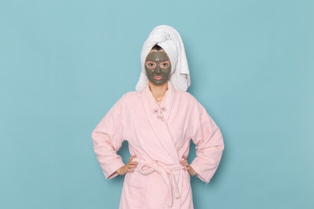 파란색 벽 샤워 청소 아름다움 셀프 케어 크림에 포즈를 취하는 그녀의 얼굴에 마스크와 분홍색 목욕 가운에 전면보기 젊은 여성