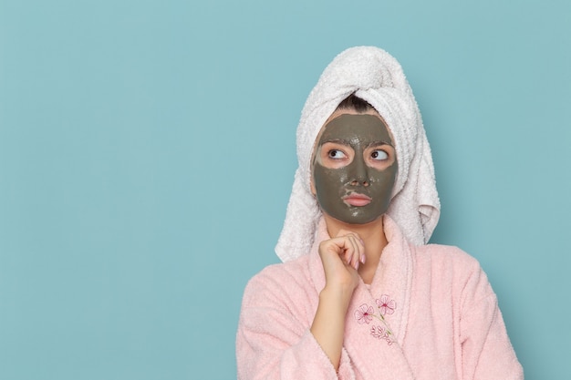 파란색 벽 샤워 청소 뷰티 셀프 케어 크림에 생각하는 그녀의 얼굴에 마스크와 분홍색 목욕 가운에 전면보기 젊은 여성
