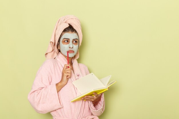 Front view young female in pink bathrobe writing down notes thinking on a green surface