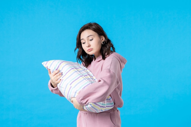 Вид спереди молодая женщина в розовой пижаме, держащая подушку на синей стене, цвет ночи, постельный отдых, эмоция, сон, бессонница, вечеринка