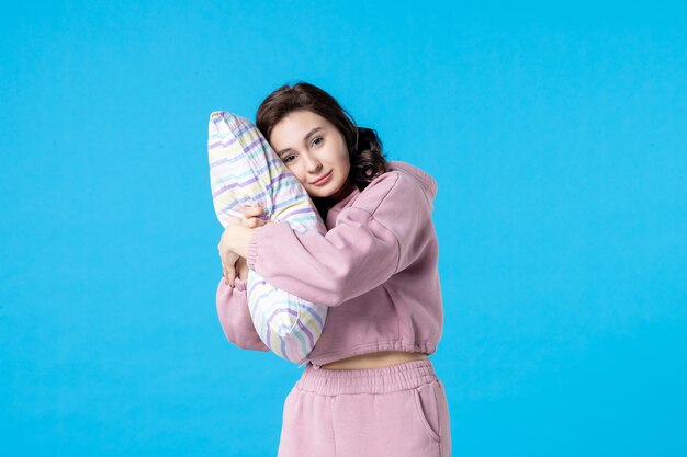 Вид спереди молодая женщина в розовой пижаме, держащая подушку на синей стене, цвет ночи, постельный отдых, эмоция, мечта, женщина, сон, бессонница