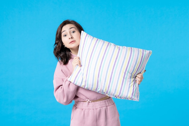 Вид спереди молодая женщина в розовой пижаме, держащая подушку на синей стене, цвет ночи, постельный отдых, эмоции, мечты, женщина, сон, бессонница