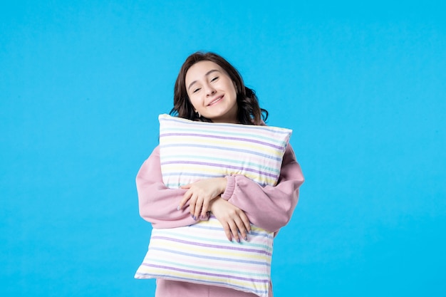 Вид спереди молодая женщина в розовой пижаме, держащая подушку на синей стене, цвет ночи, постельный отдых, модель эмоции, женщина мечты, сон