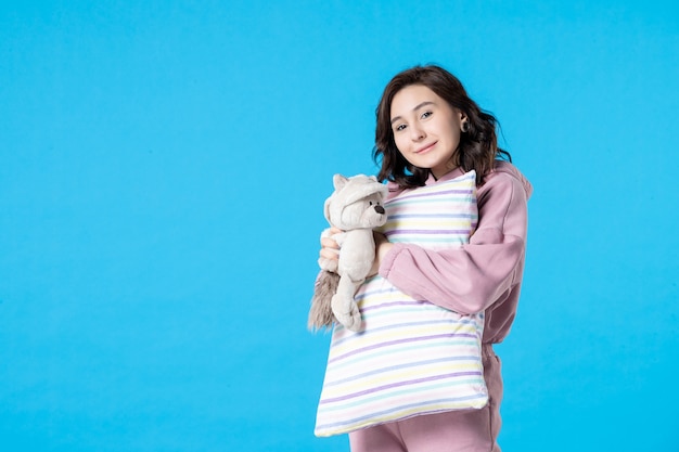 Front view young female in pink pajamas with toy bear and pillow on a light blue 