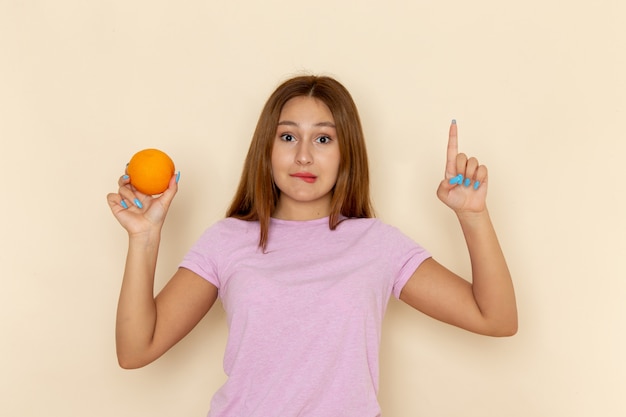 Front view young female in pink t-shirt and blue jeans holding orange with confused expression
