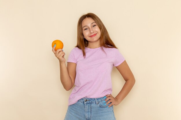 Вид спереди молодая женщина в розовой футболке и синих джинсах держит апельсин с улыбкой на лице