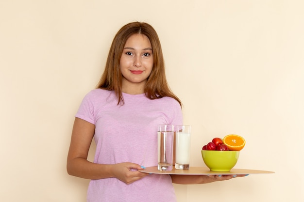 Вид спереди молодая женщина в розовой футболке и синих джинсах, держащая поднос с фруктами, молоком и водой на сером