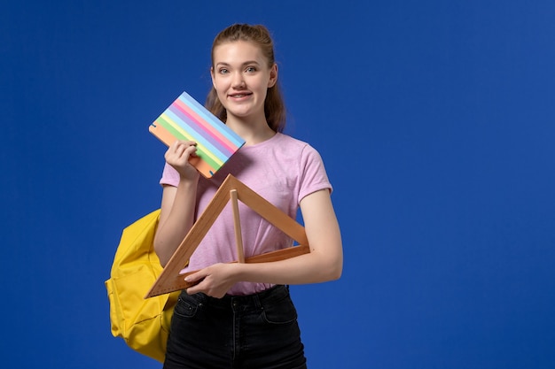 Front view of young female in pink t-shirt holding wooden triangle figure and copybook smiling on the blue wall