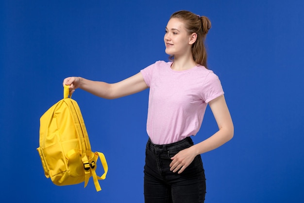 Vista frontale di giovane donna in maglietta rosa che tiene zaino giallo sulla parete blu