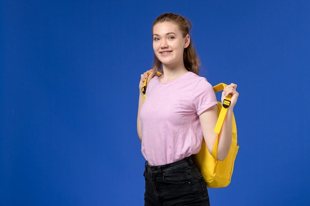Front view of young female in pink t-shirt holding yellow backpack smiling on blue wall