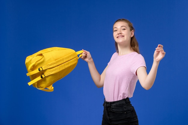 青い壁に笑みを浮かべて黄色のバックパックを保持しているピンクのTシャツの若い女性の正面図