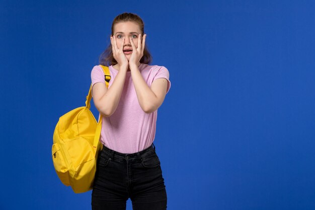 青い壁に黄色のバックパックを身に着けているピンクのTシャツの若い女性の正面図