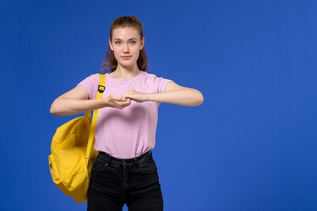 青い壁に黄色のバックパックを身に着けているピンクのTシャツの若い女性の正面図