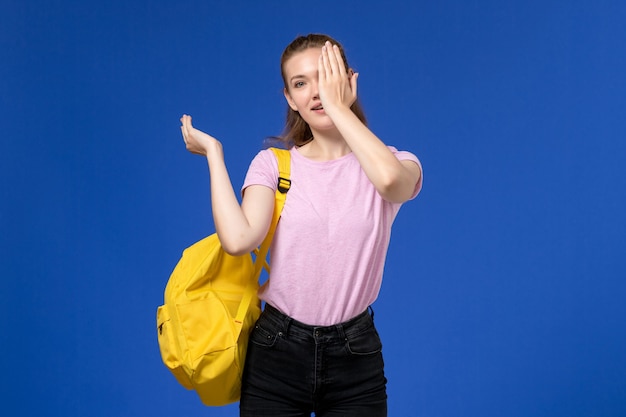 Front view of young female in pink t-shirt wearing yellow backpack closing half of her face on the light-blue wall