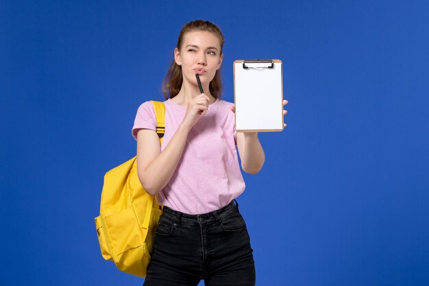 Front view of young female in pink t-shirt wearing yellow backpack and holding notepad thinking on blue wall