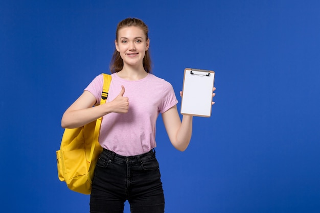 黄色のバックパックを身に着けて、青い壁に笑顔でメモ帳を保持しているピンクのTシャツの若い女性の正面図