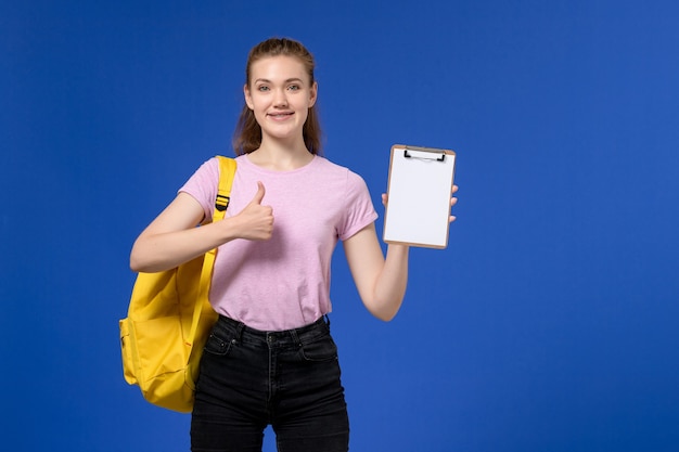 Front view of young female in pink t-shirt wearing yellow backpack and holding notepad with smile on blue wall