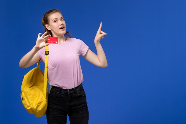 青い壁にプラスチックの赤いカードを保持している黄色のバックパックを身に着けているピンクのTシャツの若い女性の正面図