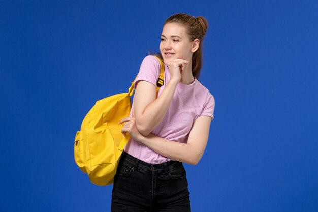 青い壁にポーズをとって黄色のバックパックを身に着けているピンクのTシャツの若い女性の正面図