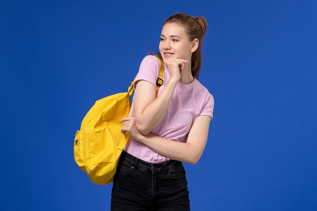 Вид спереди молодой женщины в розовой футболке с желтым рюкзаком, позирующей на синей стене