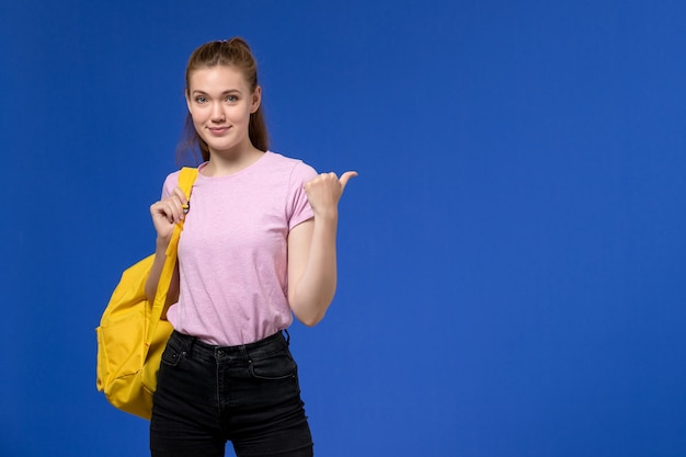 青い壁にポーズをとって黄色のバックパックを身に着けているピンクのTシャツの若い女性の正面図