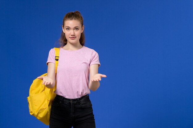Front view of young female in pink t-shirt wearing yellow backpack posing on light blue wall