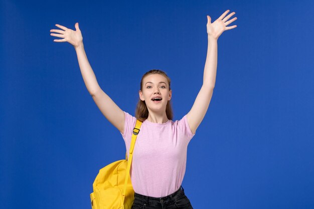 Front view of young female in pink t-shirt wearing yellow backpack raising her hands on blue wall