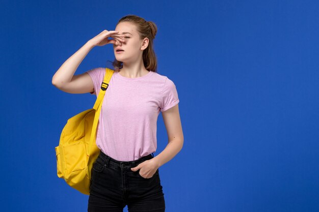 Front view of young female in pink t-shirt wearing yellow backpack shutting her nose on the light-blue wall