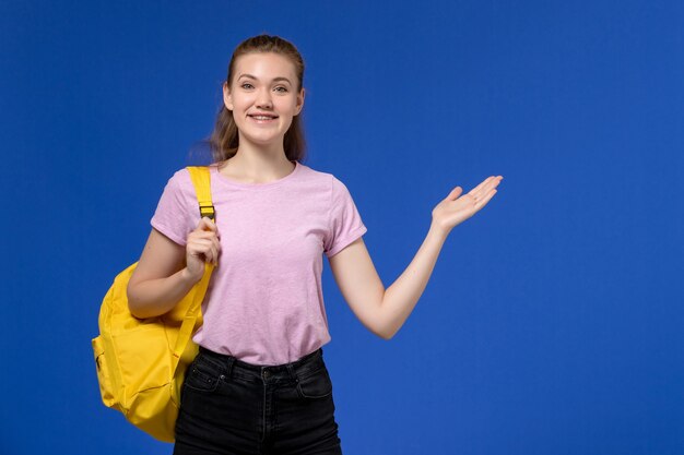 Front view of young female in pink t-shirt wearing yellow backpack smiling on light blue wall