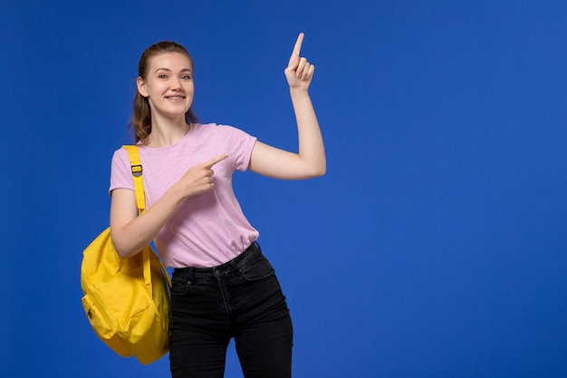 Front view of young female in pink t-shirt wearing yellow backpack smiling on light blue wall