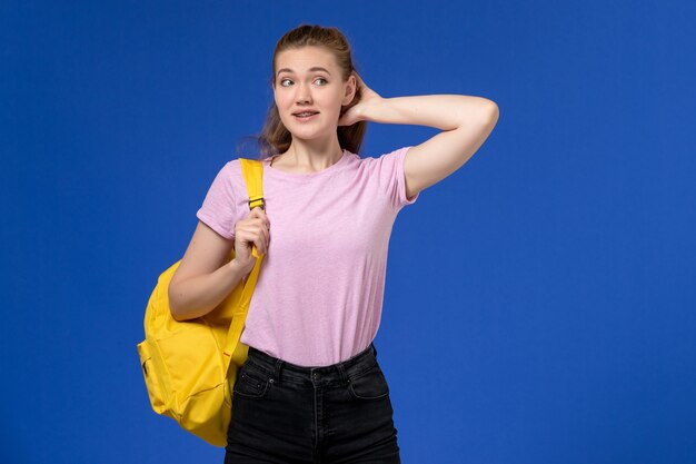 Front view of young female in pink t-shirt wearing yellow backpack thinking on the light-blue wall
