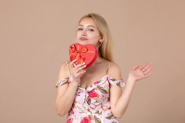 Vista frontale di una giovane donna in posa con un regalo a forma di cuore rosso sul muro marrone