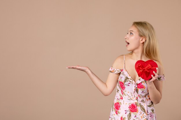 Foto gratuita vista frontale di una giovane donna in posa con un regalo a forma di cuore rosso sul muro marrone