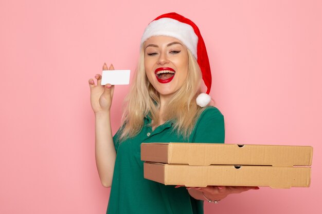Vista frontale della giovane donna con berretto rosso in possesso di carta di credito e scatole per pizza sulla parete rosa
