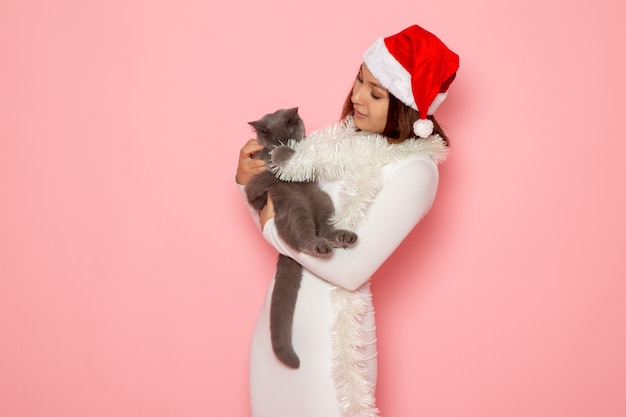 Front view of young female in red cap holding cute grey kitten on pink wall