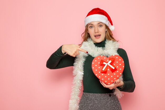 Front view of young female in red cap holding heart shaped present on pink wall