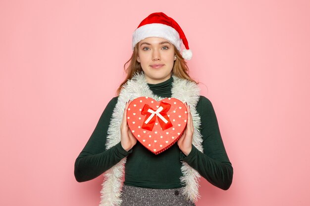 Front view of young female in red cap holding heart shaped present on pink wall