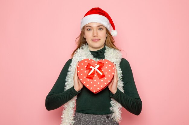 Вид спереди молодой женщины в красной кепке, держащей подарок в форме сердца на розовой стене