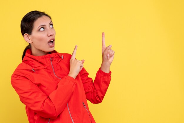 Front view young female in red coat pointing at ceiling on yellow background