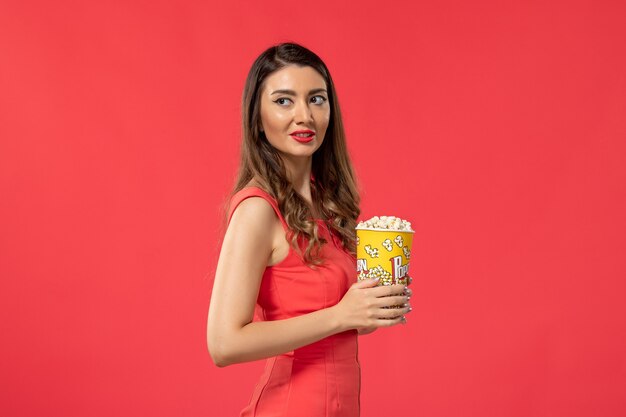 Вид спереди молодая женщина в красной рубашке, держащая попкорн, смотрит фильм на красной поверхности