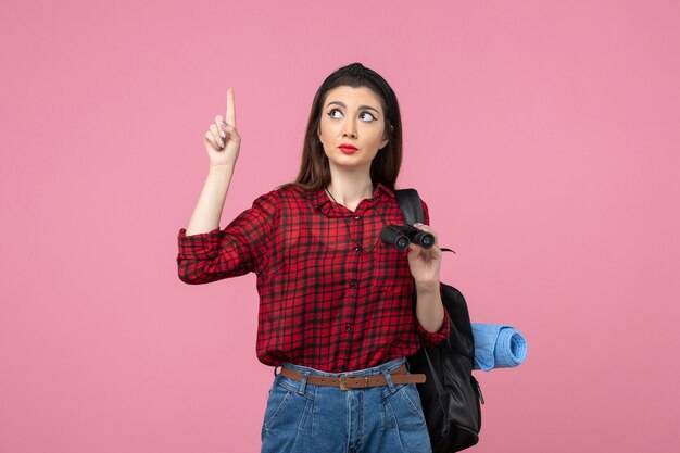 Front view young female in red shirt with binoculars on the pink background woman color student