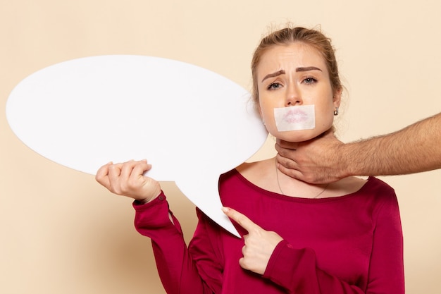 Front view young female in red shirt with tied mouth holding white sign with man hands around her neck on the cream space female cloth  photo