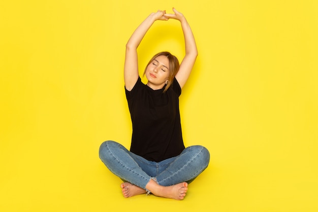 Free photo a front view young female sitting in black shirt and blue jeans doing gymnastics on yellow